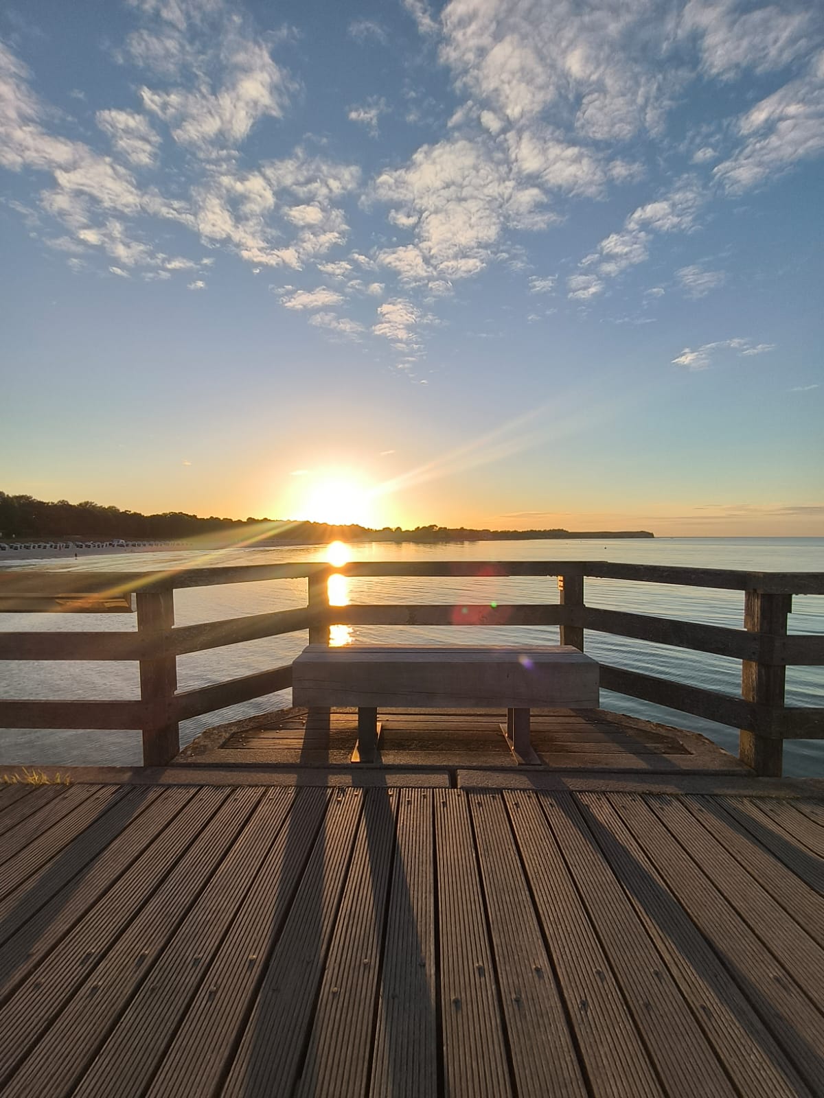 Holzsteg an der Ostsee mit Geländer und einer leeren Bank, dahinter die tief stehende Sonne über dem Wasser